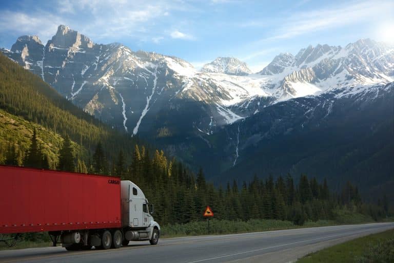 Ein roter Sattelschlepper, der Güter transportiert, fährt eine Straße entlang, im Hintergrund Berge.