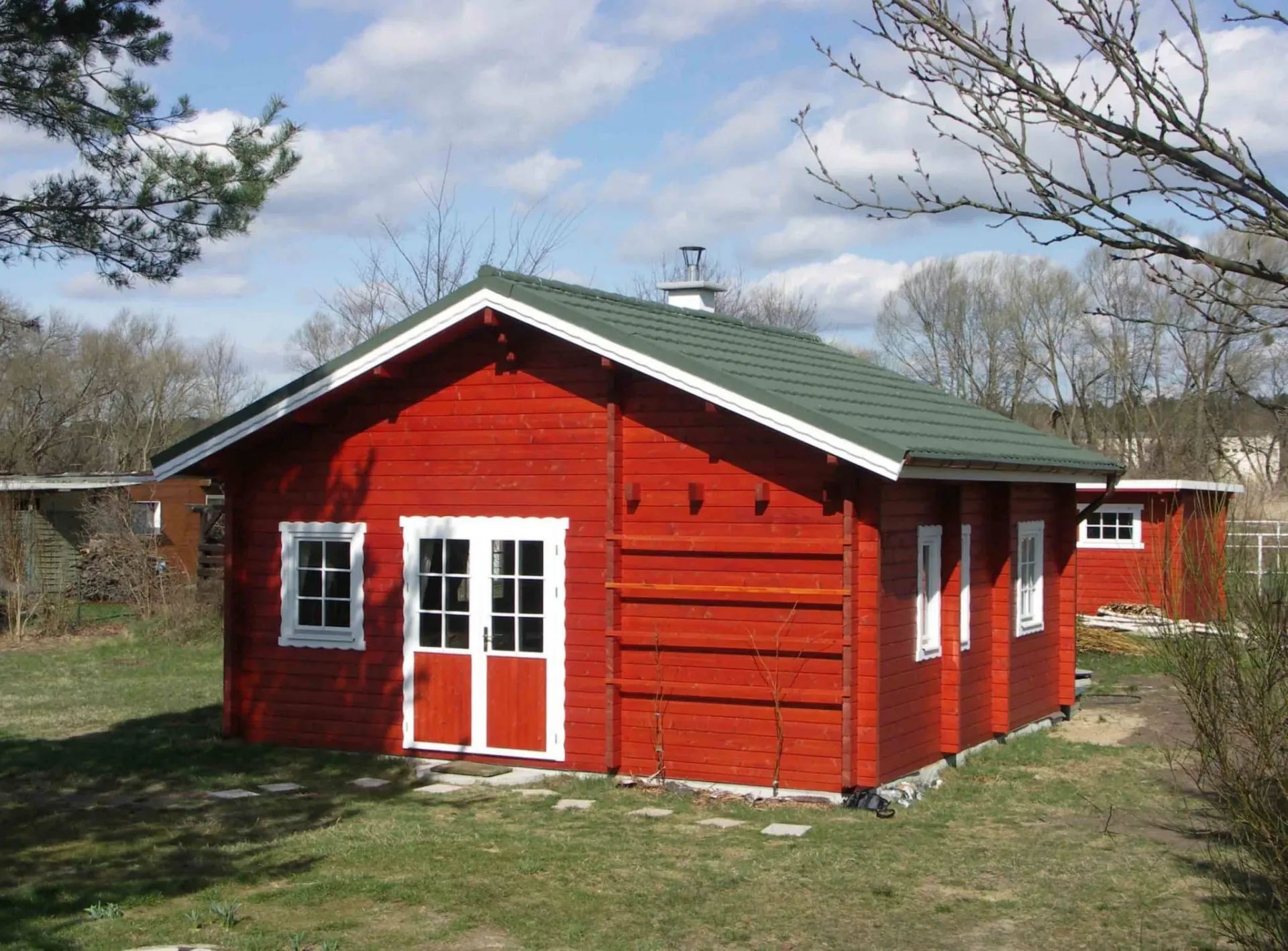 Rotes Gartenhaus aus Holz mit weißen Fenstern und grünem Dach auf einer Wiese, im Frühjahr